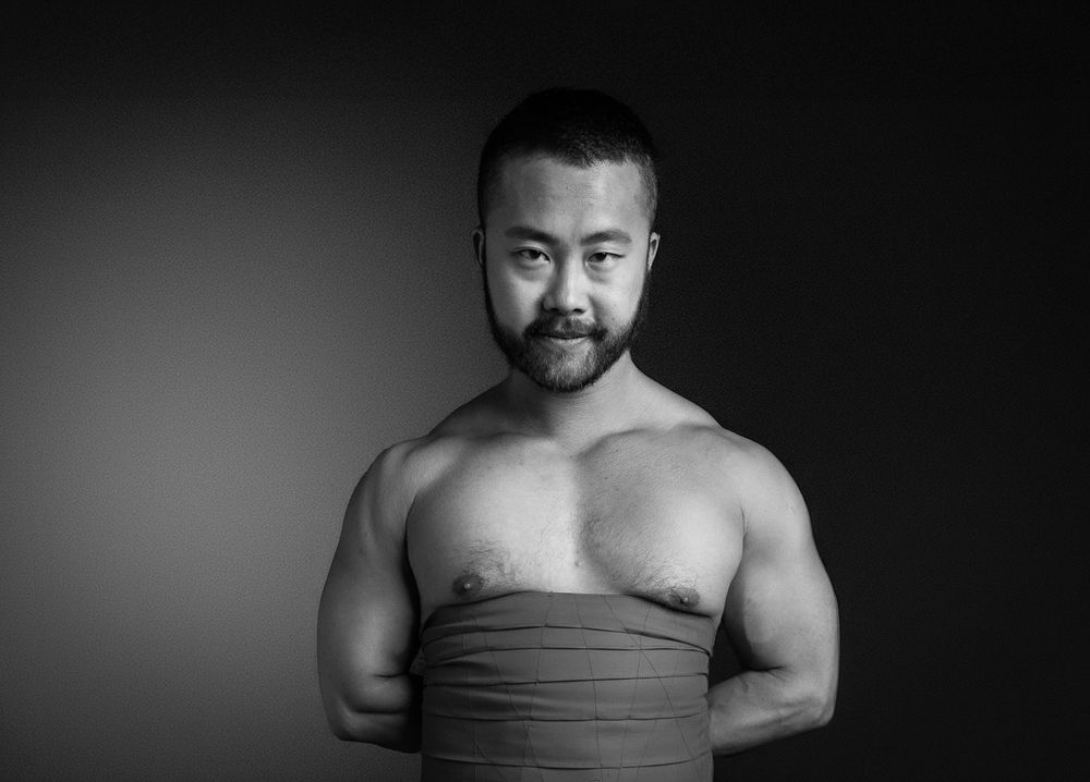 Black and white portrait of a shirtless muscular Asian man wrapped in fabric, studio background.
