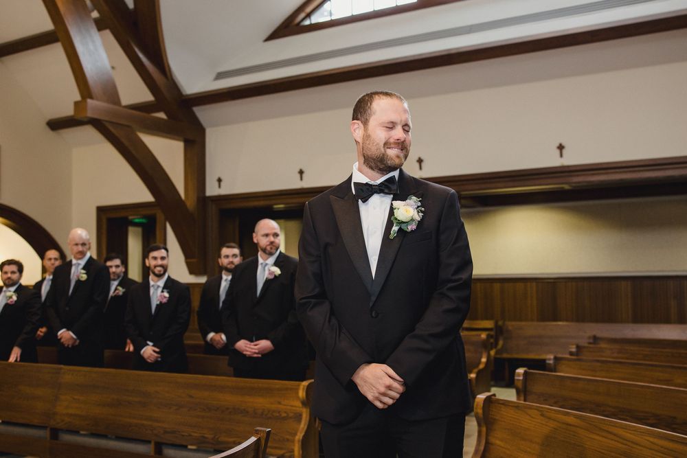 Groomsmen in black tuxedos stand in a row inside a traditional church sanctuary with wooden pews.