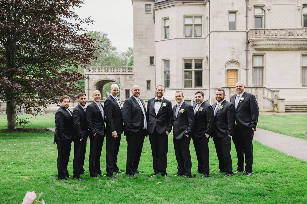 Wedding party members in black suits pose together on a lush green lawn outside an elegant stone building.