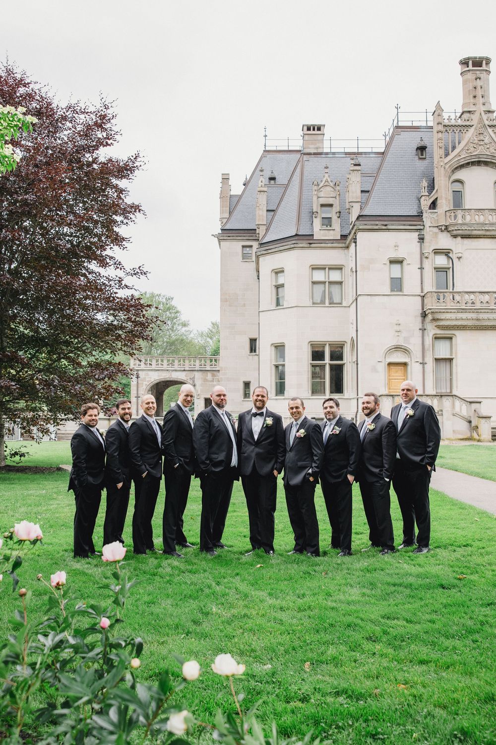 A line of groomsmen in black tuxedos stand on a green lawn in front of a grand stone mansion.