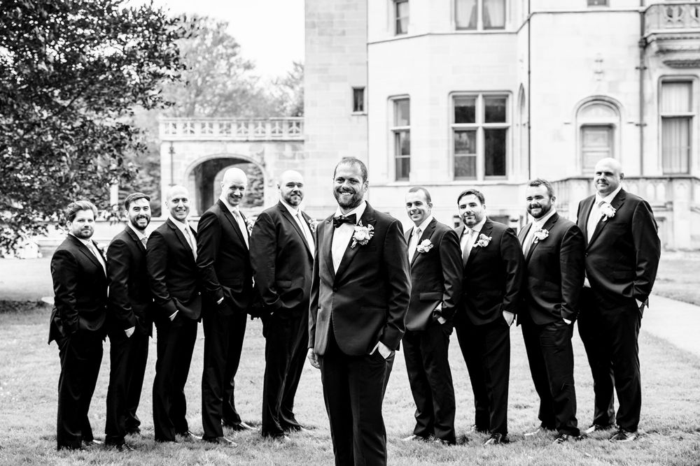 Wedding party members in formal black attire pose together in a line outdoors on a sunny day.