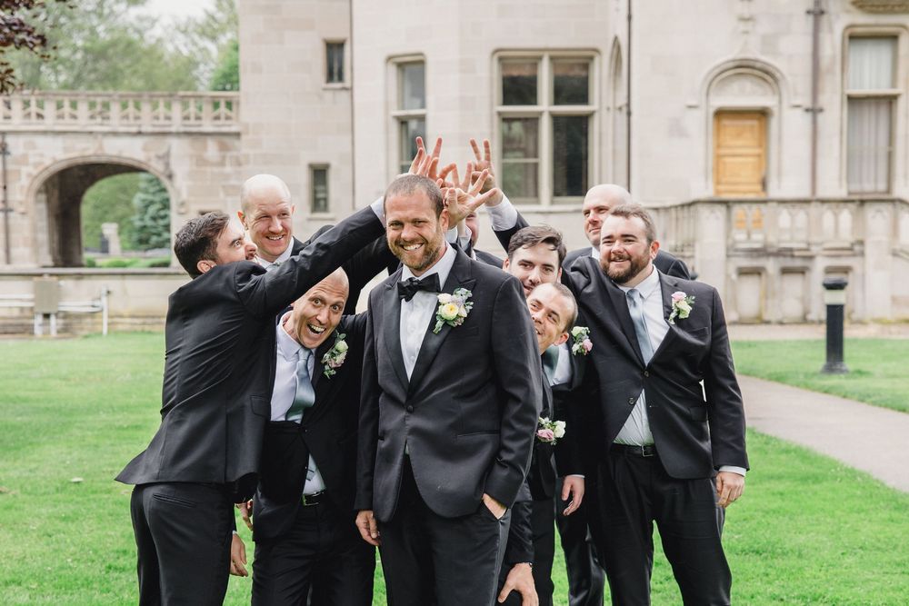 A wedding party in black tuxedos and formal attire poses together on the lawn of an elegant stone building.