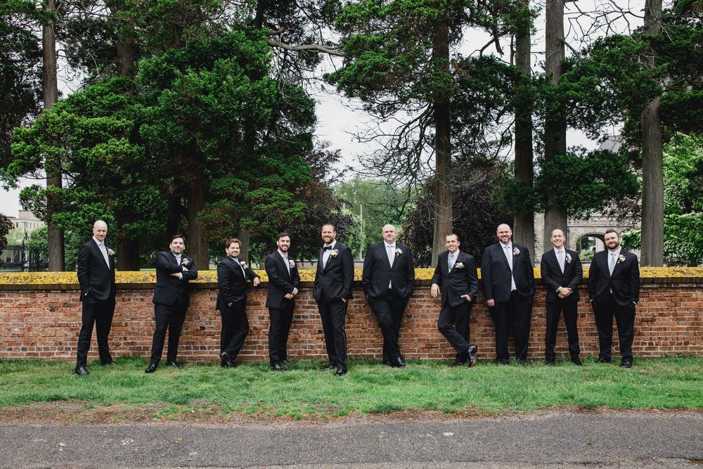 A wedding party in formal black attire stands in a line against a rustic brick wall with green trees in the background.