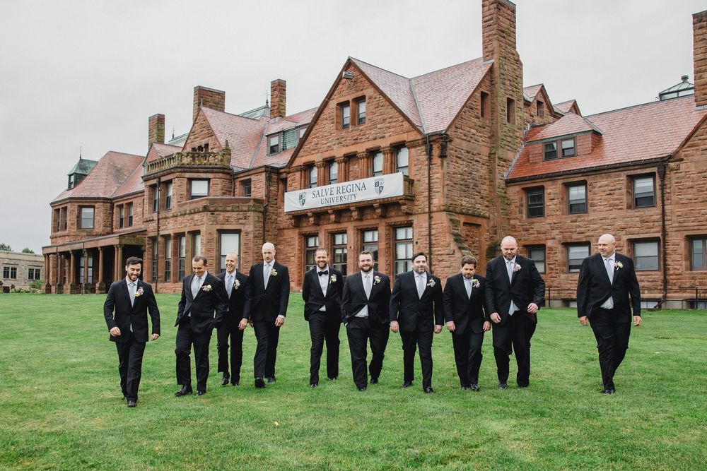 Wedding party in formal attire stands on green lawn in front of historic brick buildings at college campus.
