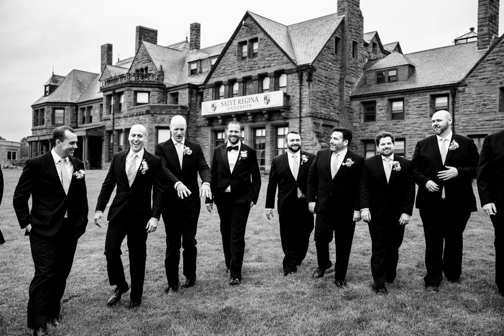 A large group of men in formal black tuxedos stand in a line against a backdrop of historic stone buildings.