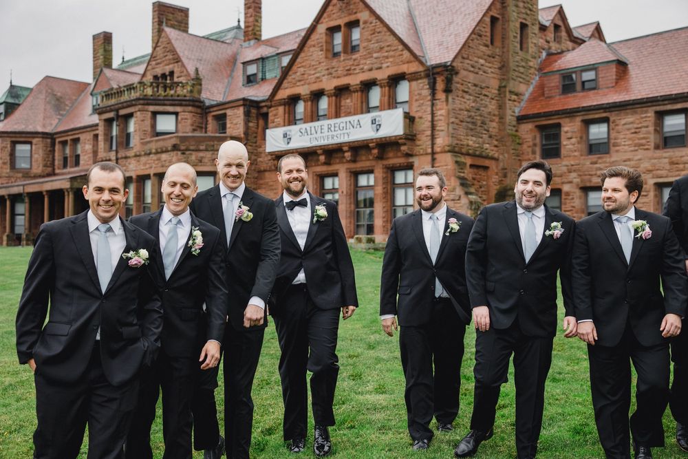 A group of groomsmen in black tuxedos stand together on a lawn in front of a grand brick mansion.
