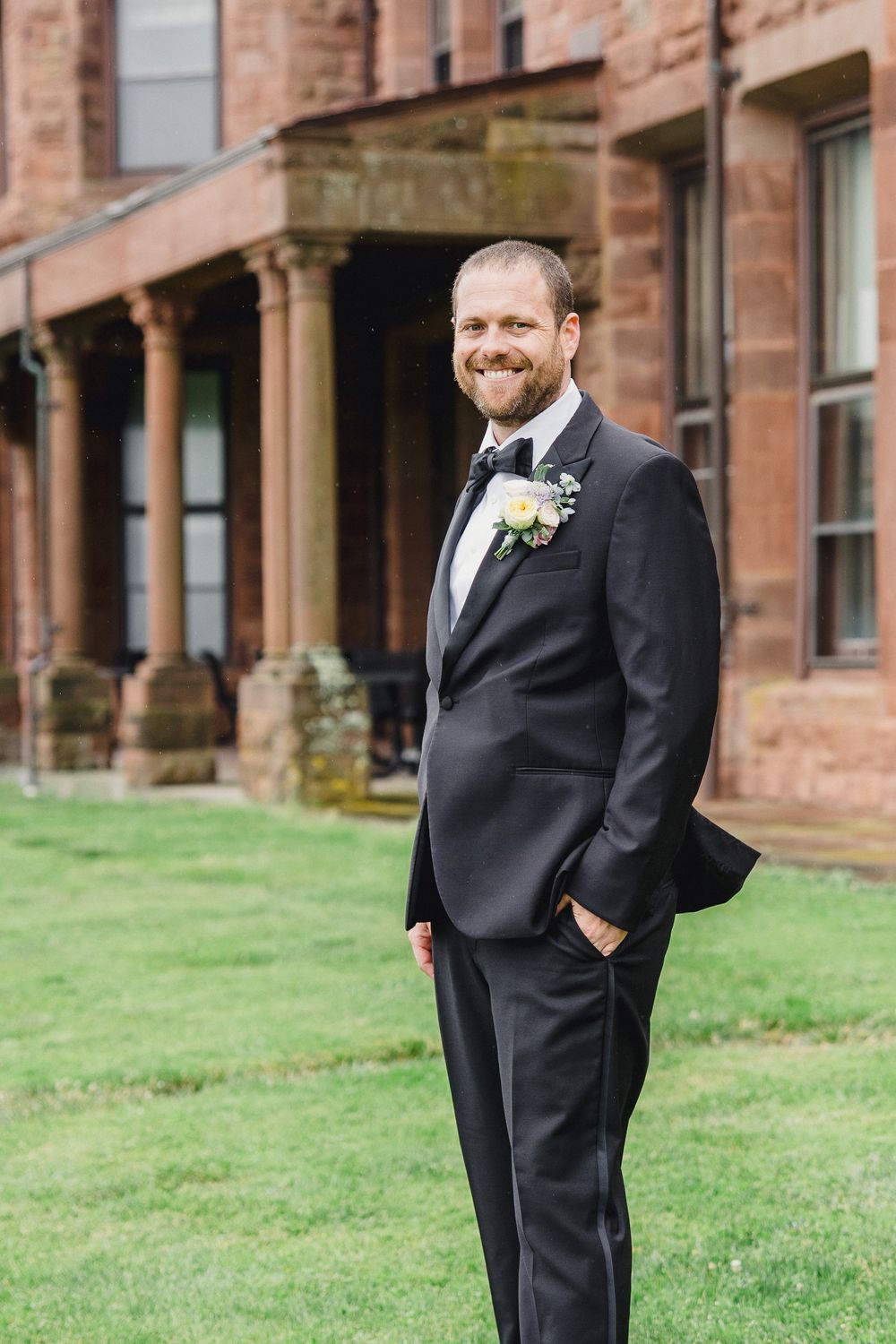 A groom in a classic black suit with boutonniere smiles while standing on a lawn outside a historic stone building.