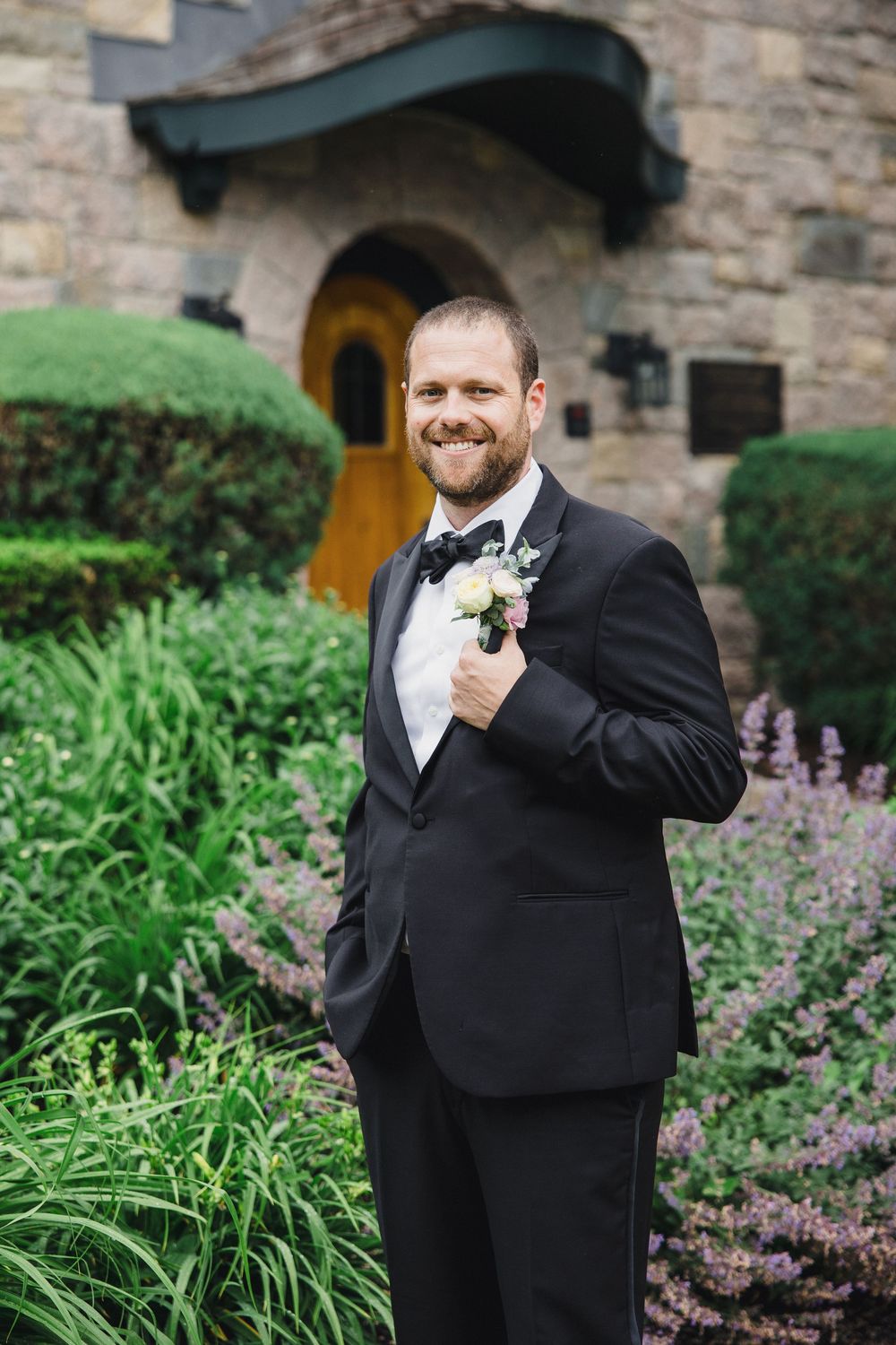 A groom in a black tuxedo smiles while adjusting his boutonniere in a garden setting with stone walls and flowers.
