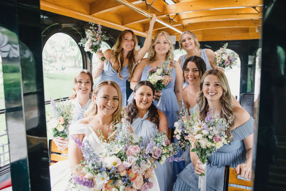 Bridesmaids in dusty blue dresses pose together holding colorful floral bouquets at a rustic wedding venue.