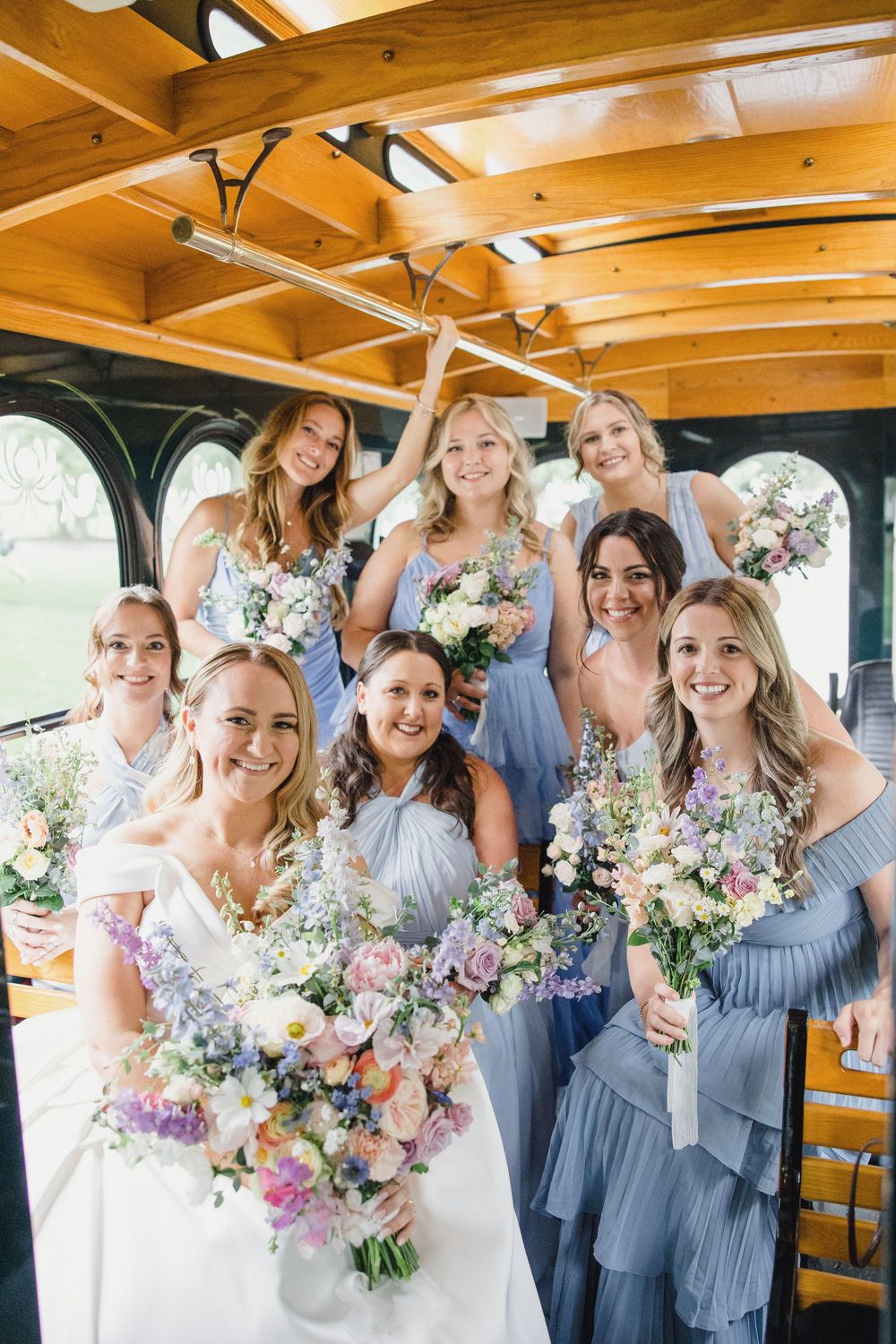 Wedding party in light blue dresses poses together holding pastel bouquets under wooden beams.