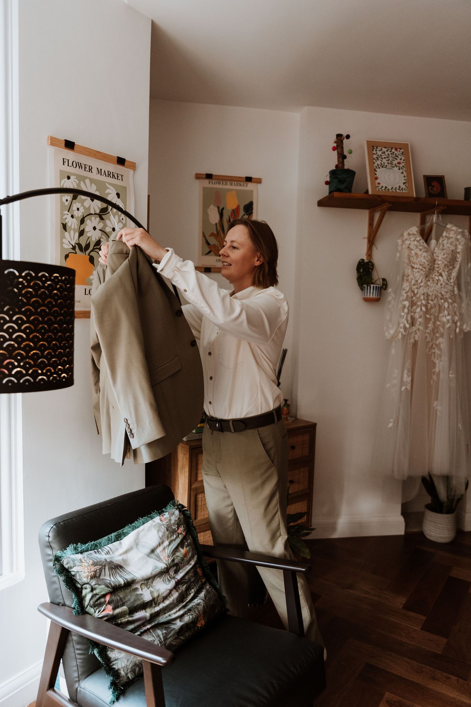 A person in white clothing hangs garments in a cozy room with wooden floors and decorative shelves displaying a wedding dress.
