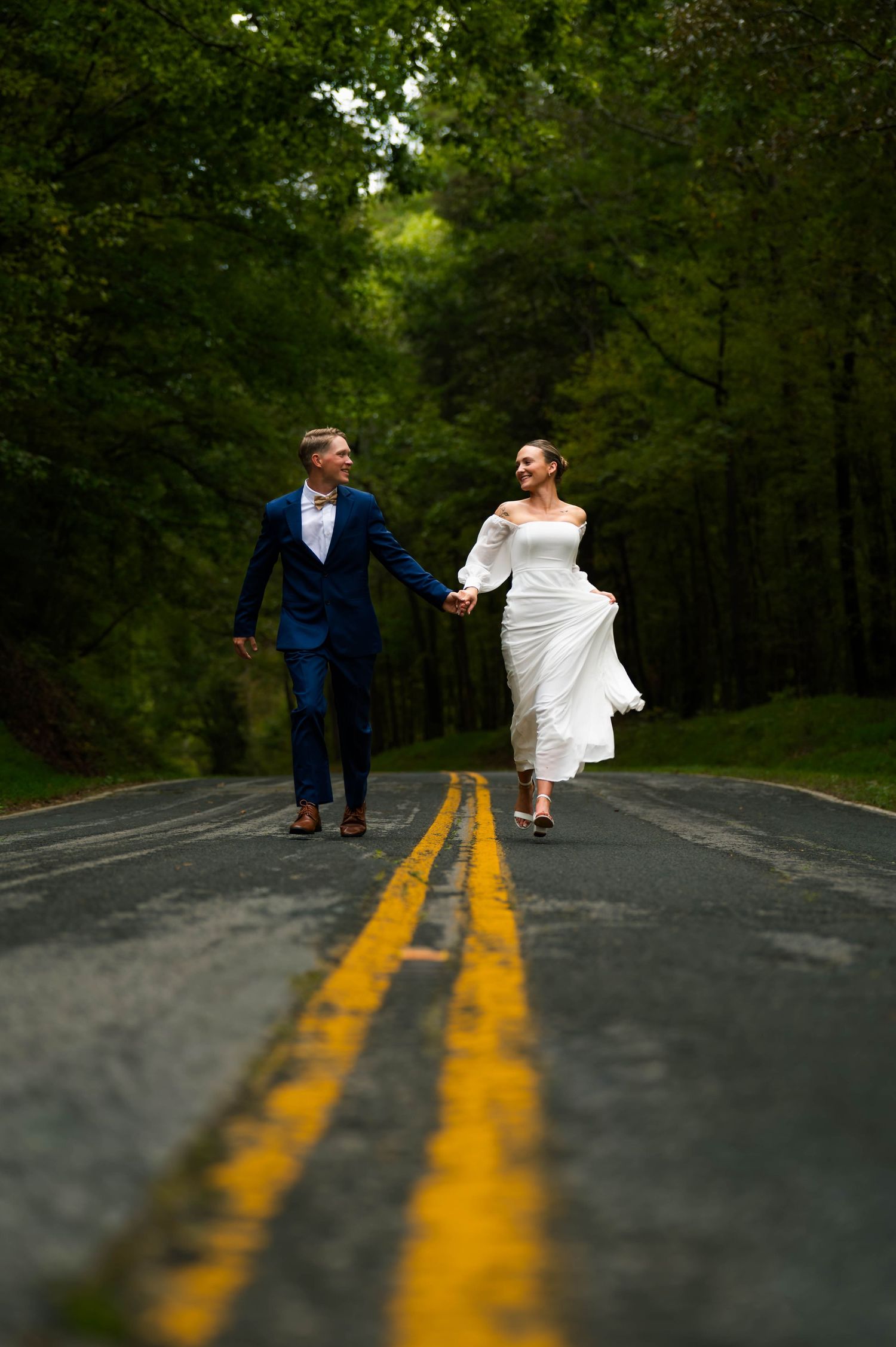 A bride and groom hold hands while running down a tree-lined road with yellow double lines, in Peak, South Carolina. Midlands SC