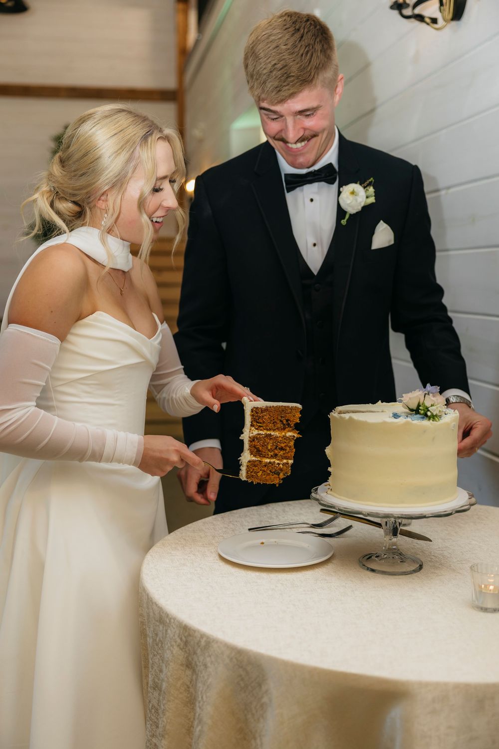 A couple in formal wedding attire share a moment of laughter while cutting their simple white wedding cake together.