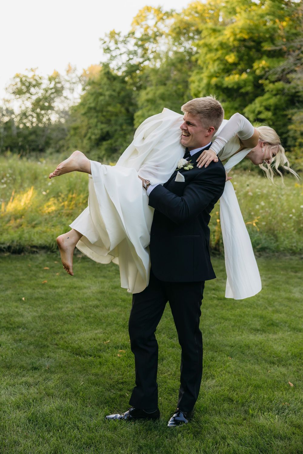 A couple in wedding attire dips playfully on a grassy hillside during a sunset photo session.