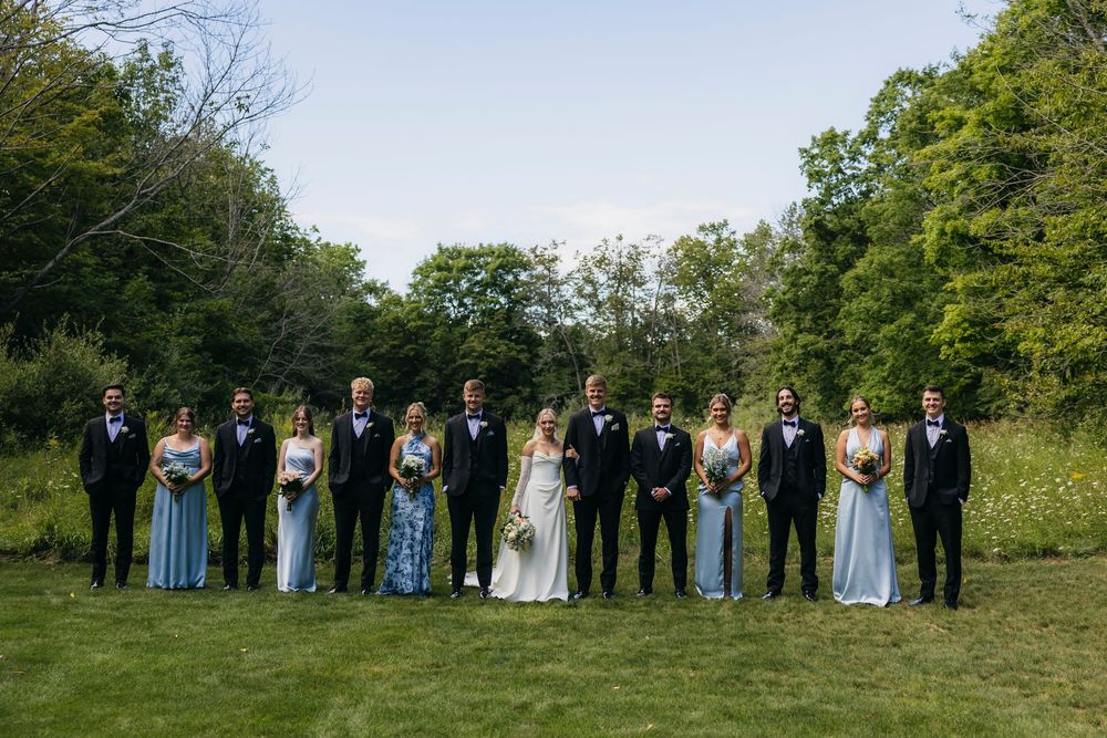Wedding party in formal attire stands in a line on green grass with lush trees in the background.