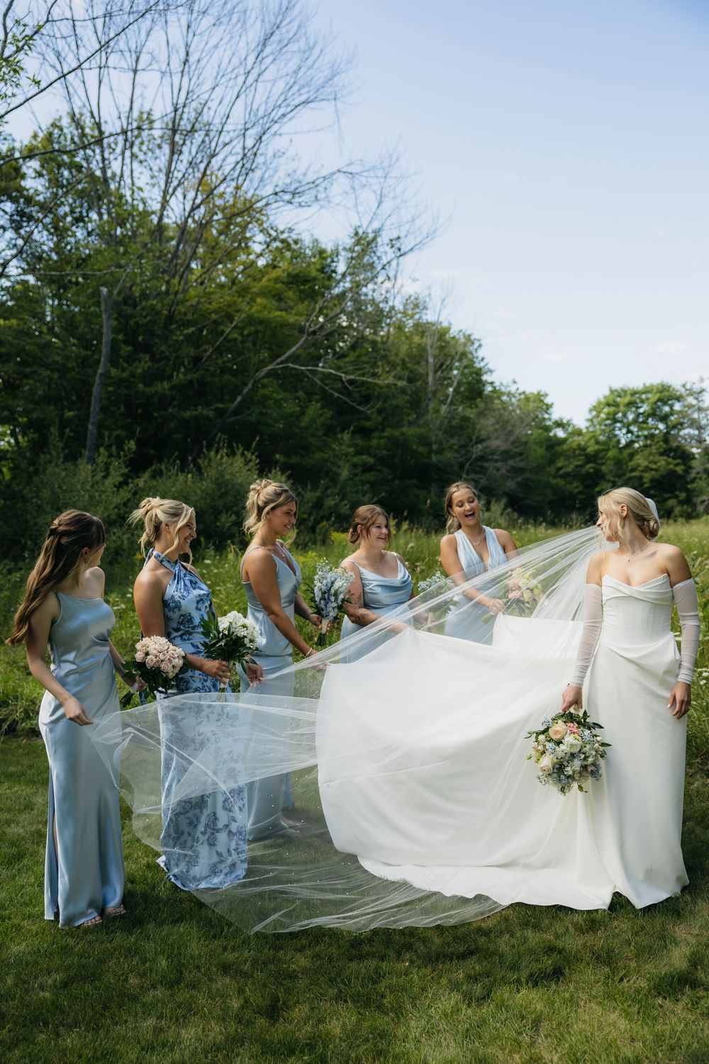 A wedding party stands on a grassy lawn as bridesmaids help adjust the bride's flowing white veil in the afternoon light.