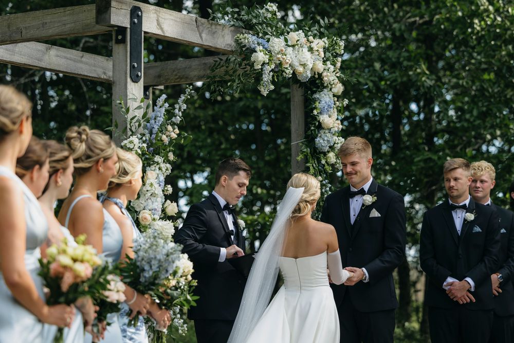 Wedding party poses outdoors in formal attire with bridesmaids in blue dresses holding white bouquets.