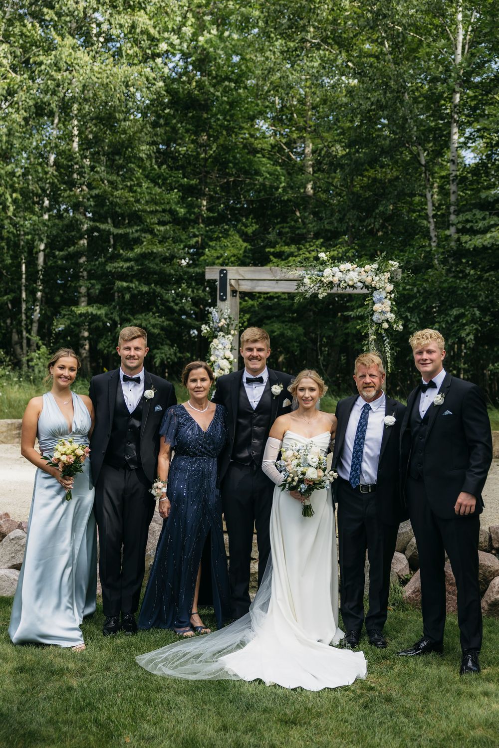Wedding party in formal attire poses together on grassy lawn with green forest backdrop.