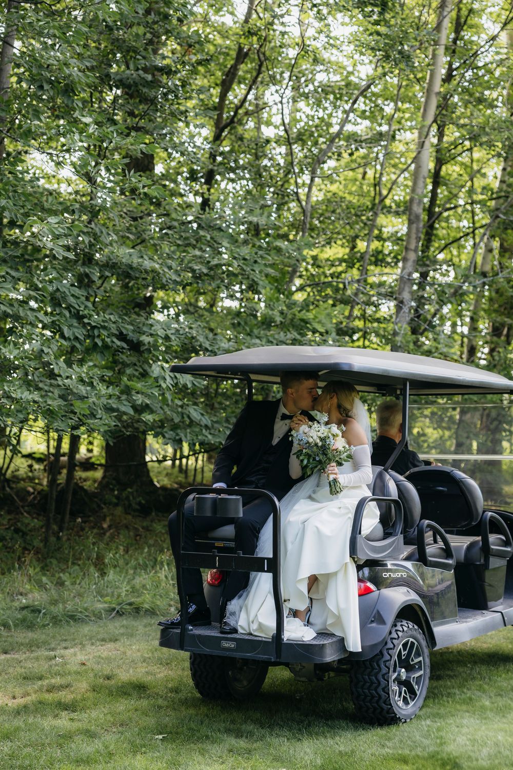 A black car parked in a grassy wooded area with people standing near its open trunk.