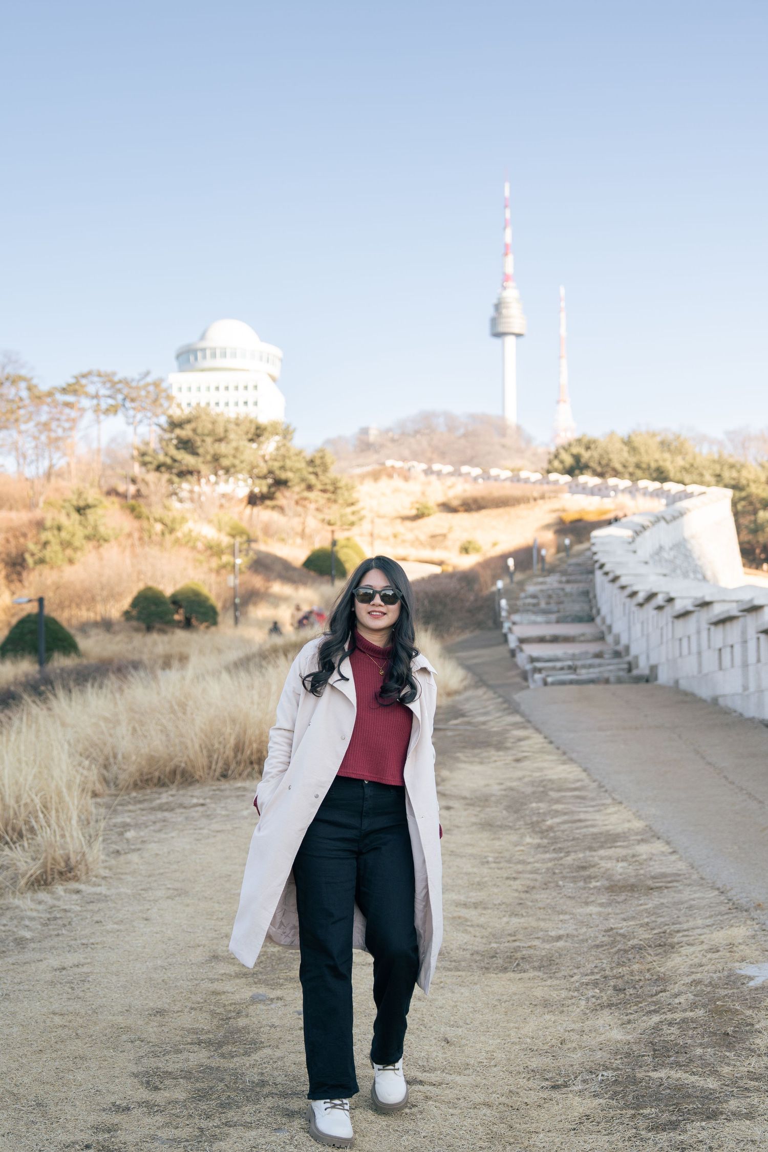 A person in a white coat and sunglasses poses along a scenic outdoor path on a sunny winter day.