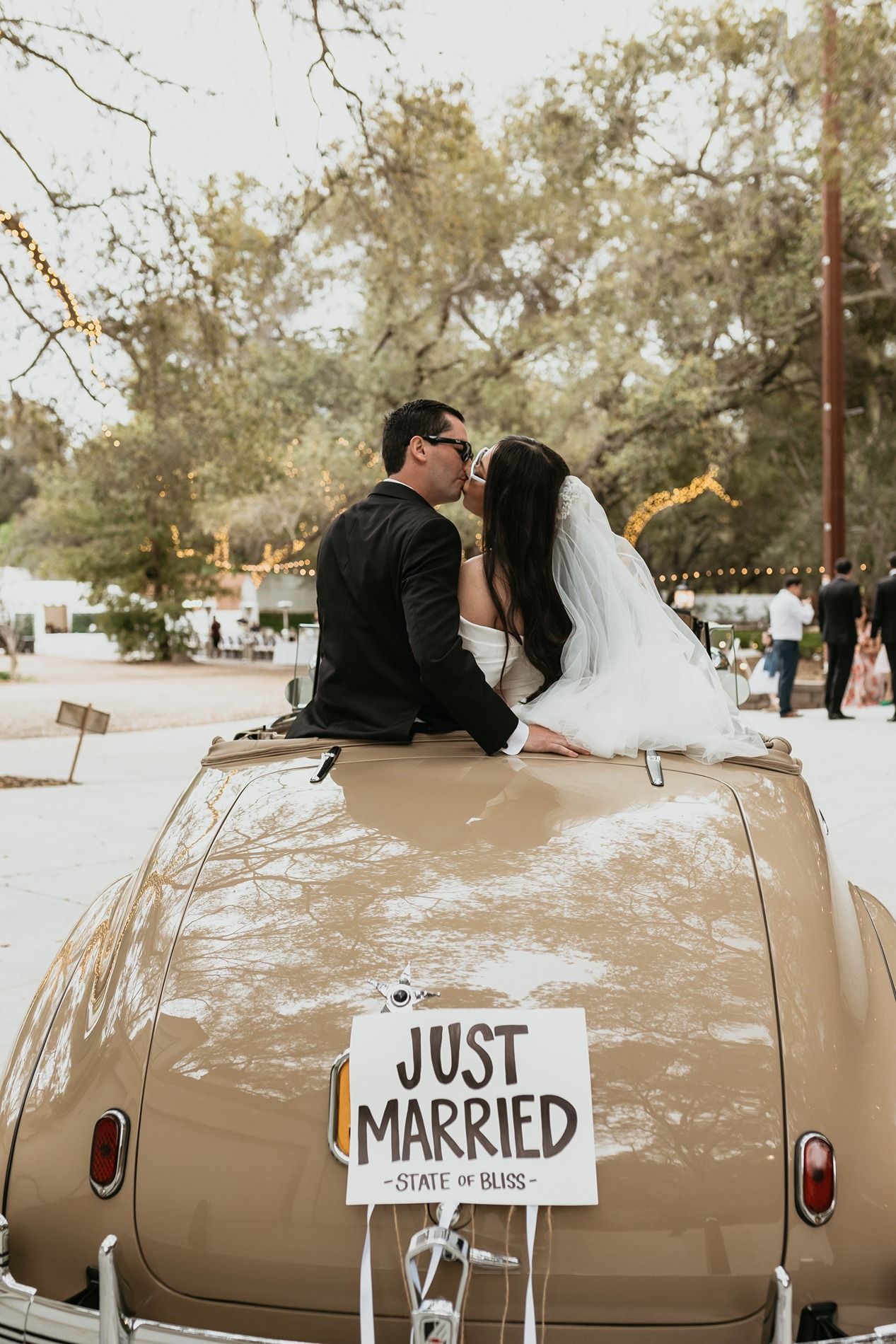A newlywed couple sits on their vintage beige car with a Just Married sign during their wedding day celebration at The Oaks at Duncan Lane.
