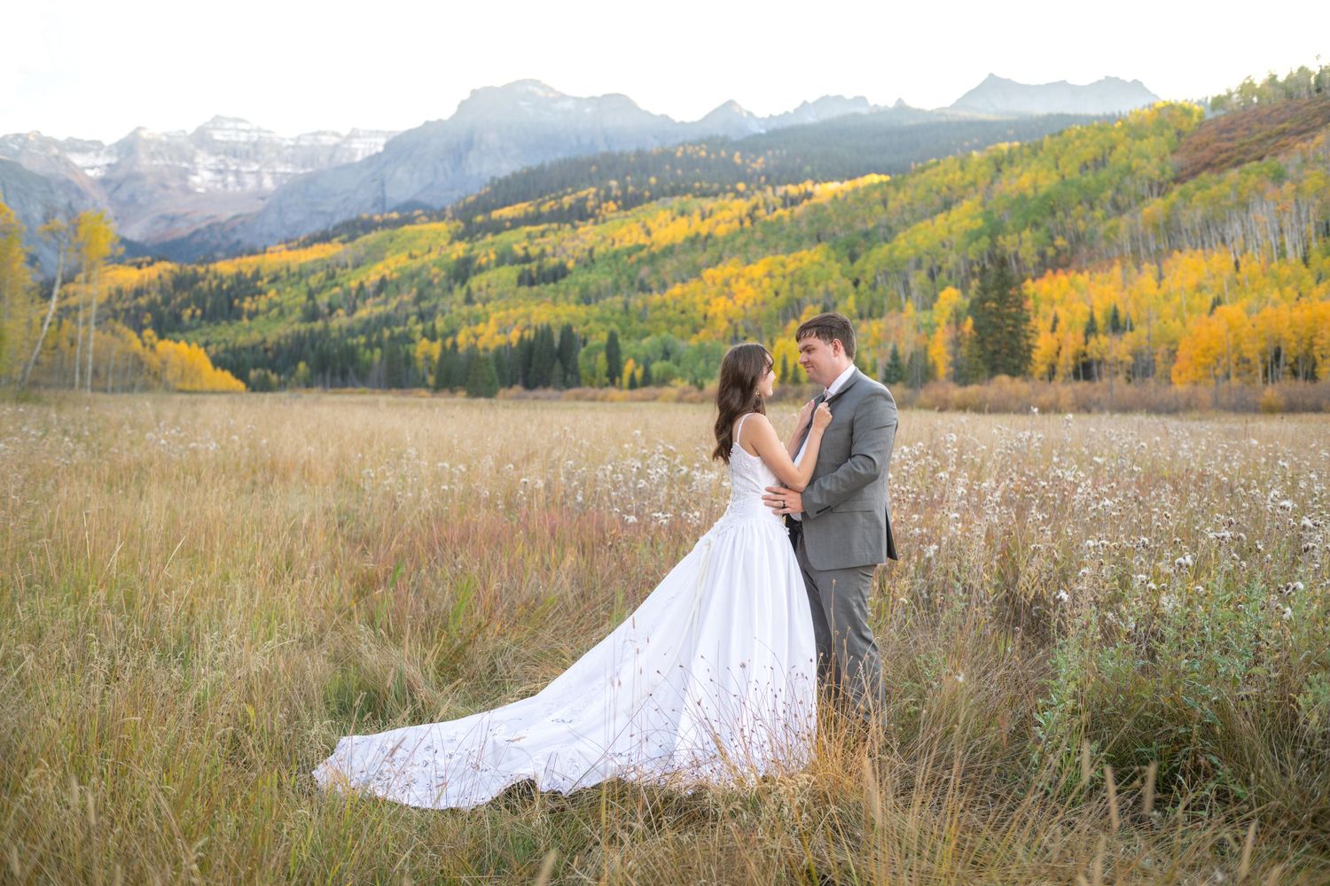 Bride and groom embrace in golden autumn meadow with aspen trees and rugged mountain peaks in background.