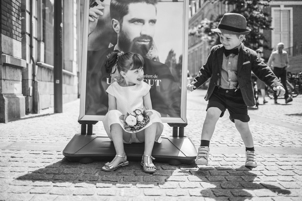 Two children in vintage clothing pose near a storefront on a cobblestone street in black and white.