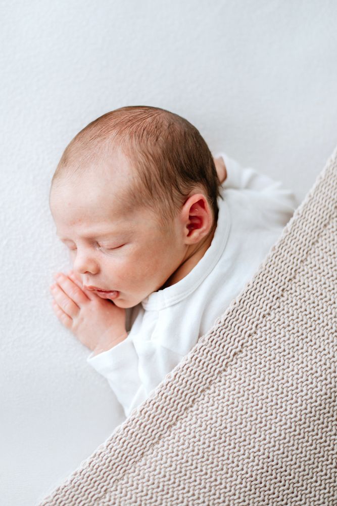A sleeping newborn baby swaddled in white rests peacefully draped in a textured beige blanket.