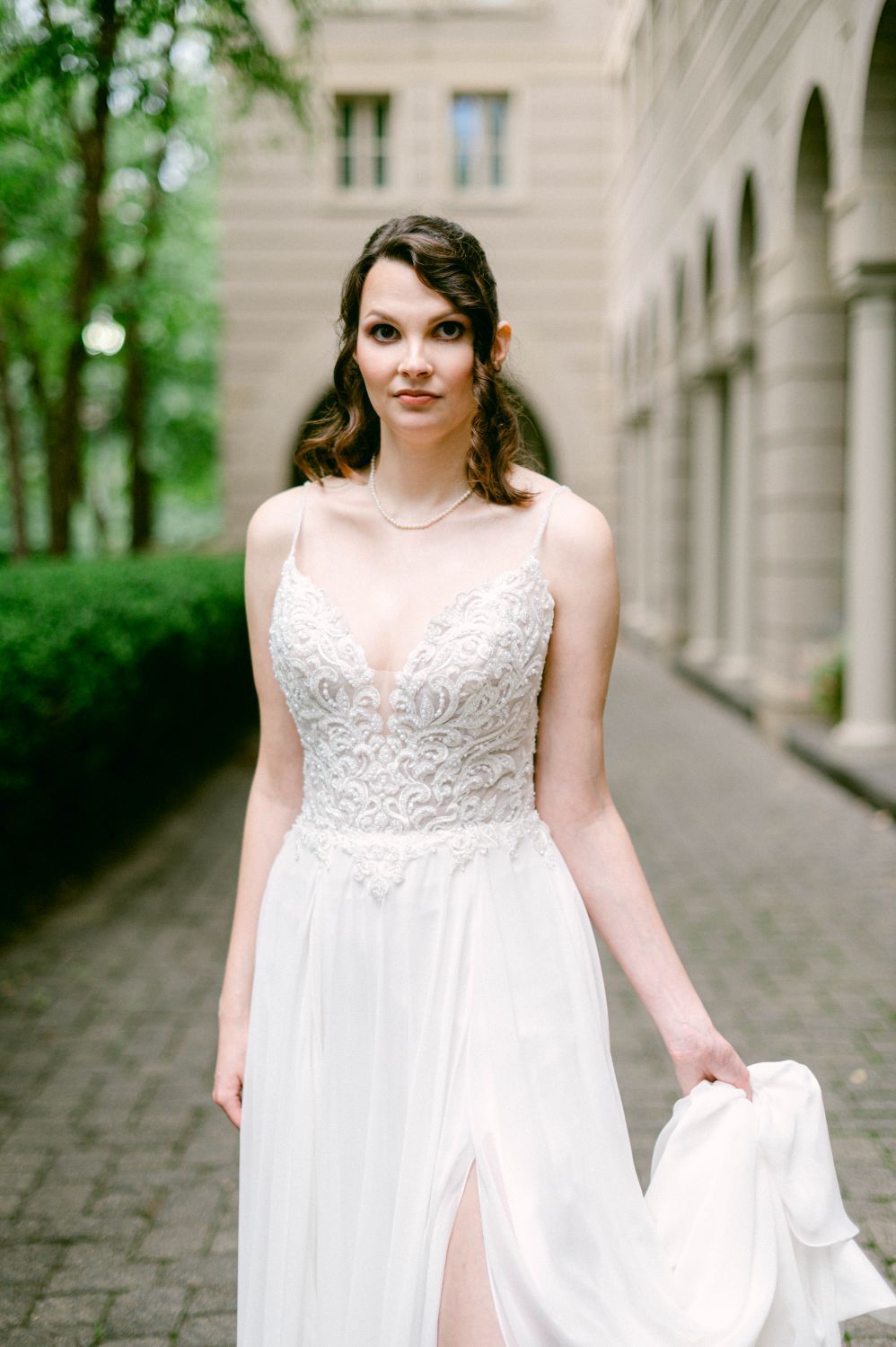 A bride in a flowing white dress with lace bodice poses on a stone-paved walkway surrounded by greenery.