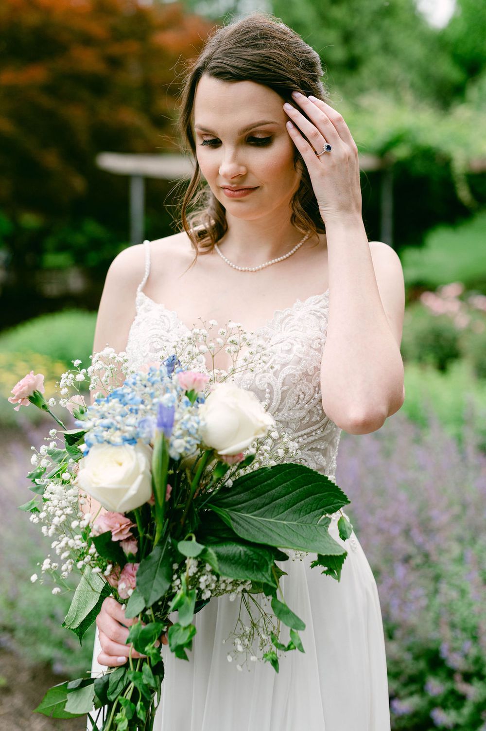A bride in a lace wedding dress holds a delicate bouquet of flowers in a garden with purple blooms in the background.