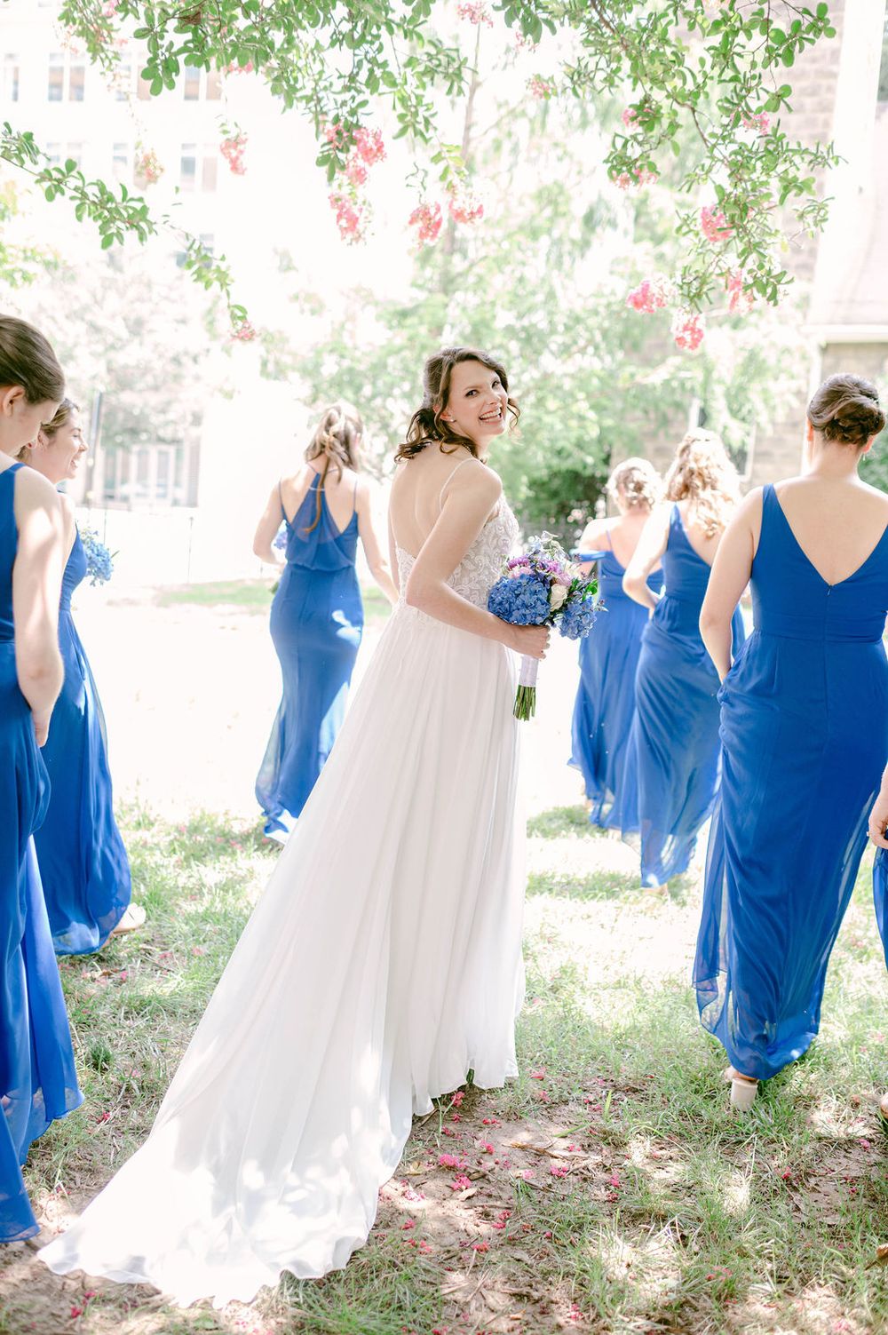 A wedding party wearing royal blue bridesmaid dresses and white bridal gowns pose together outdoors under trees.