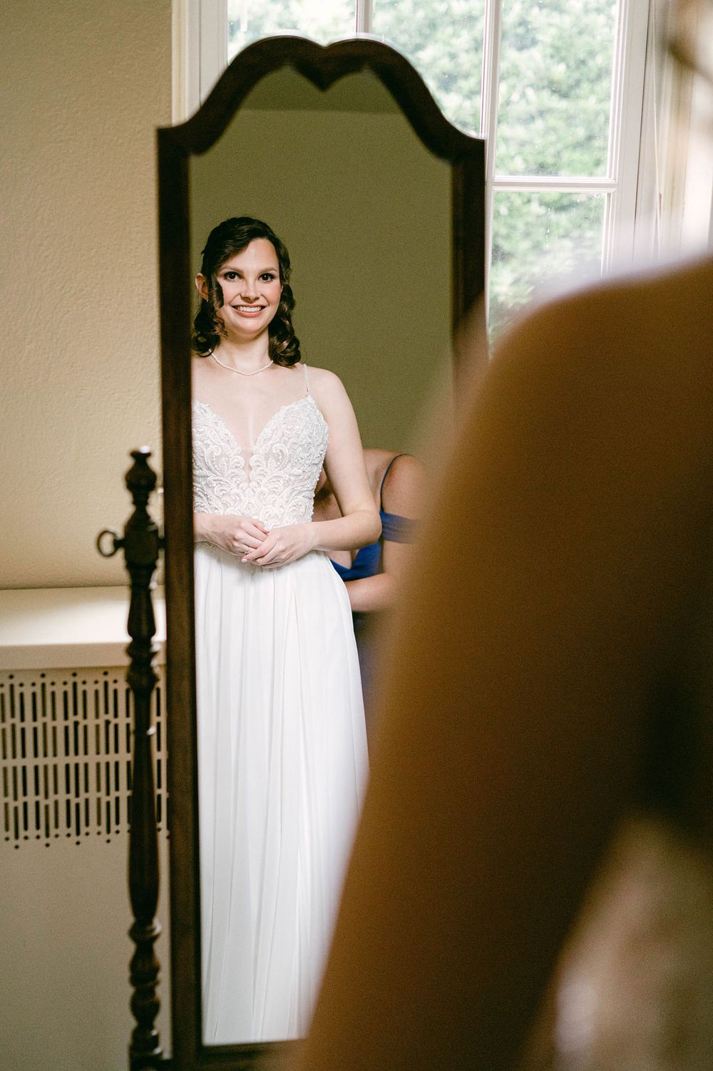 A bride in a flowing white wedding dress with lace details stands in a softly lit room with vintage decor.
