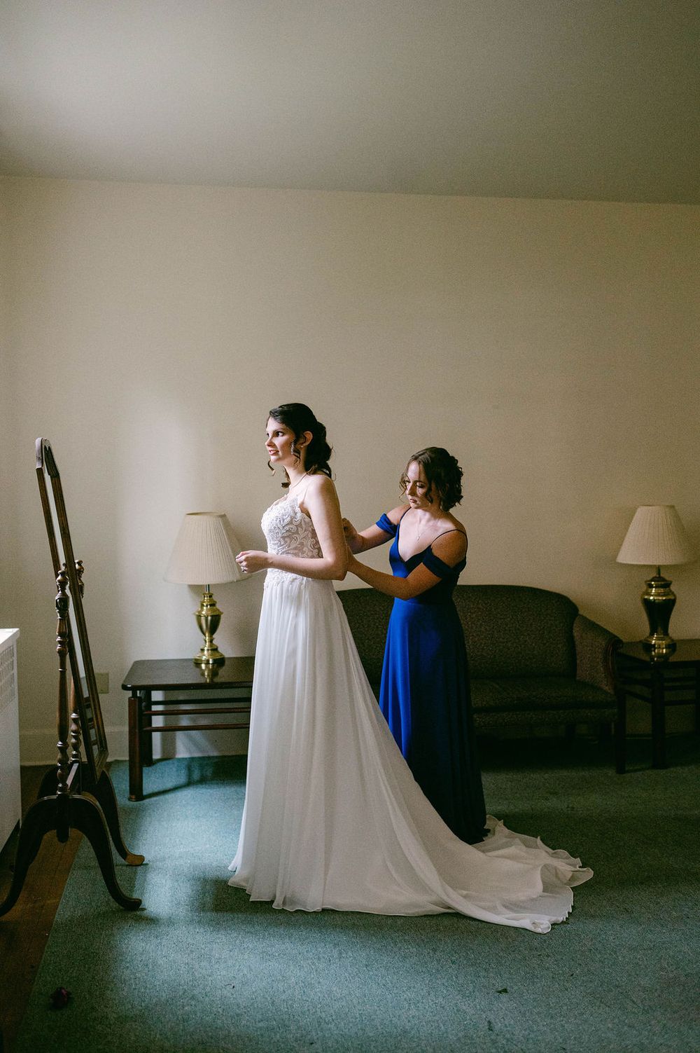 Two people in formal attire prepare for a special occasion, with one wearing a flowing white gown and another in royal blue.