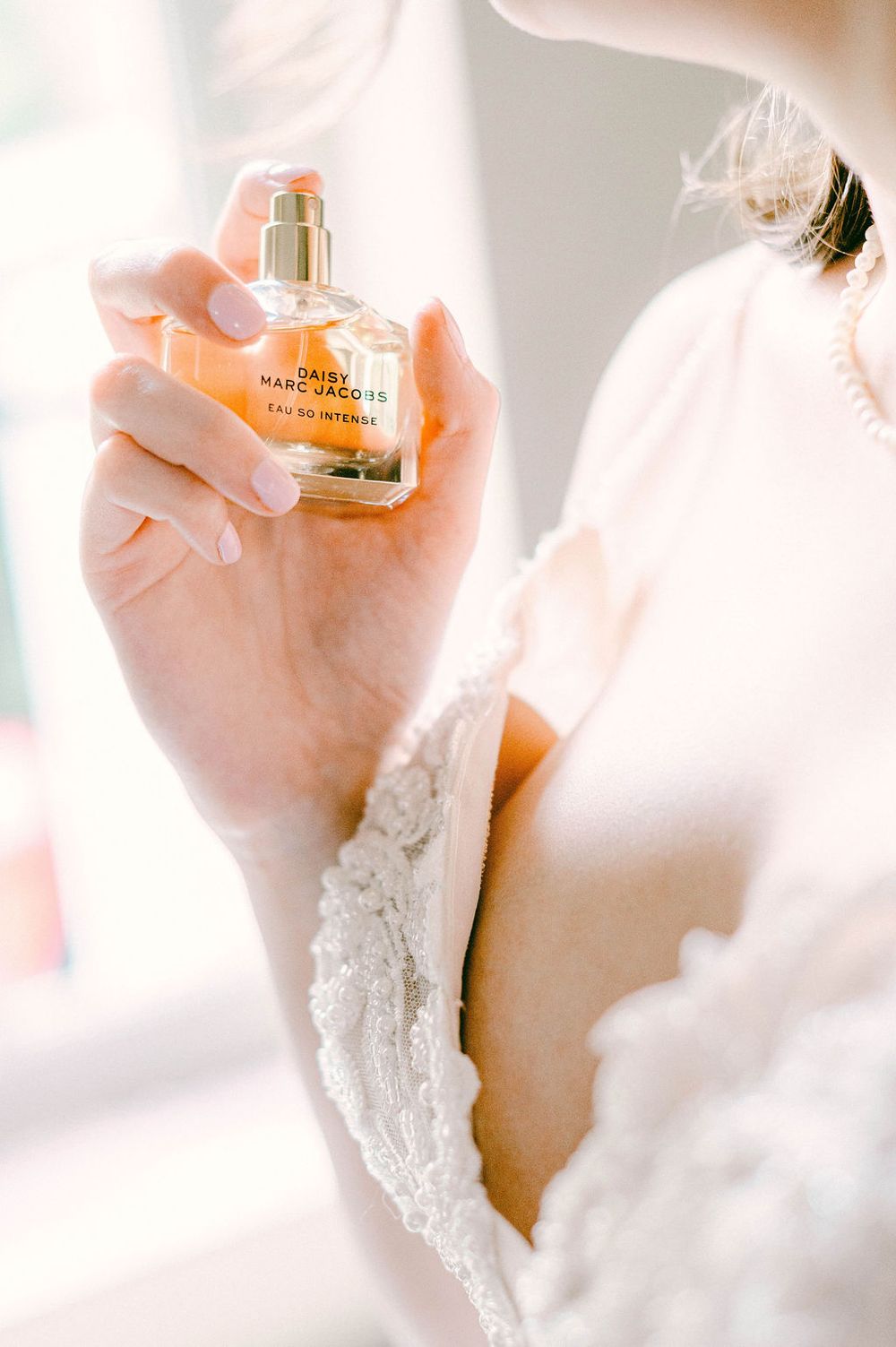 A delicate hand holding a perfume bottle against a soft white lace background in dreamy lighting.