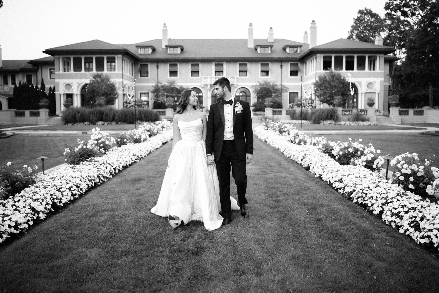 Wedding couple poses in front of elegant building with white floral arrangements lining the walkway.