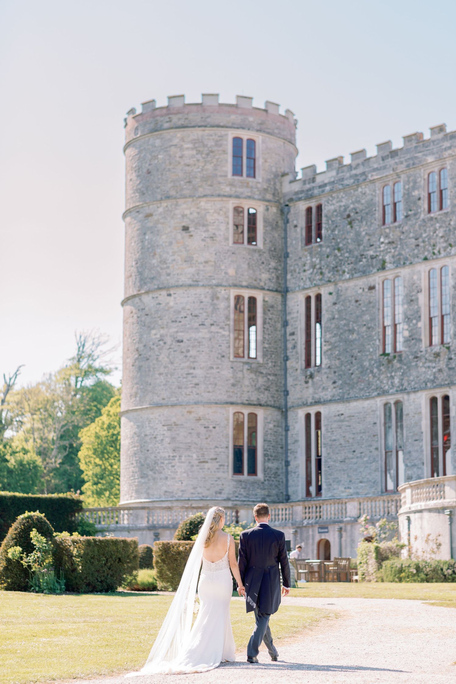 A couple walks hand in hand on a path leading to a grand stone castle with a circular tower on a sunny day.