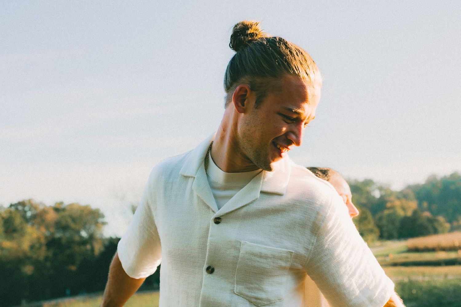 Person in white button-up shirt walking through sunny field at golden hour with lens flare effect.