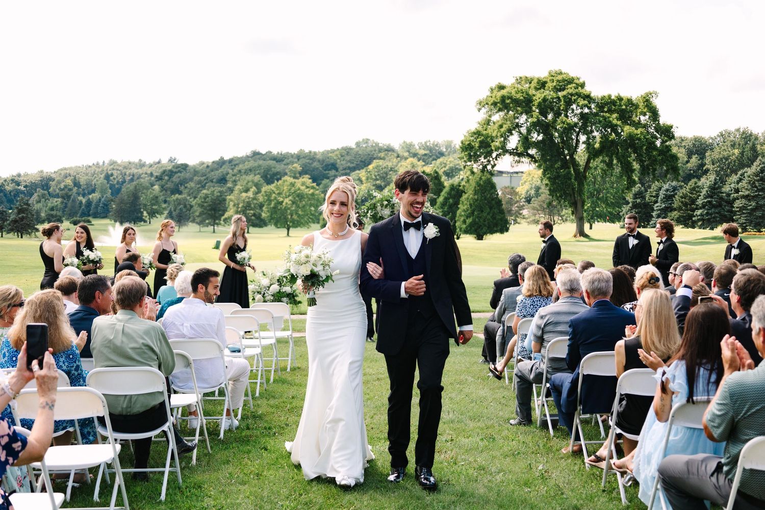Panoramic view of an outdoor wedding ceremony with guests seated in white chairs on a grassy hillside.