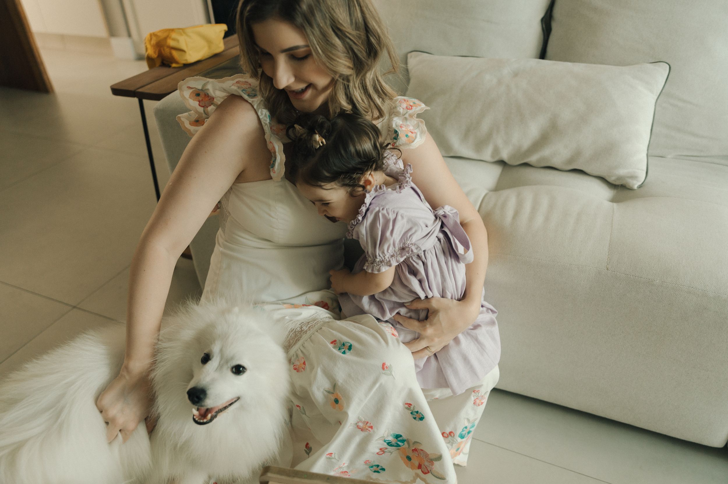 A person sits on a beige couch with a fluffy white dog and young child in a pink dress.