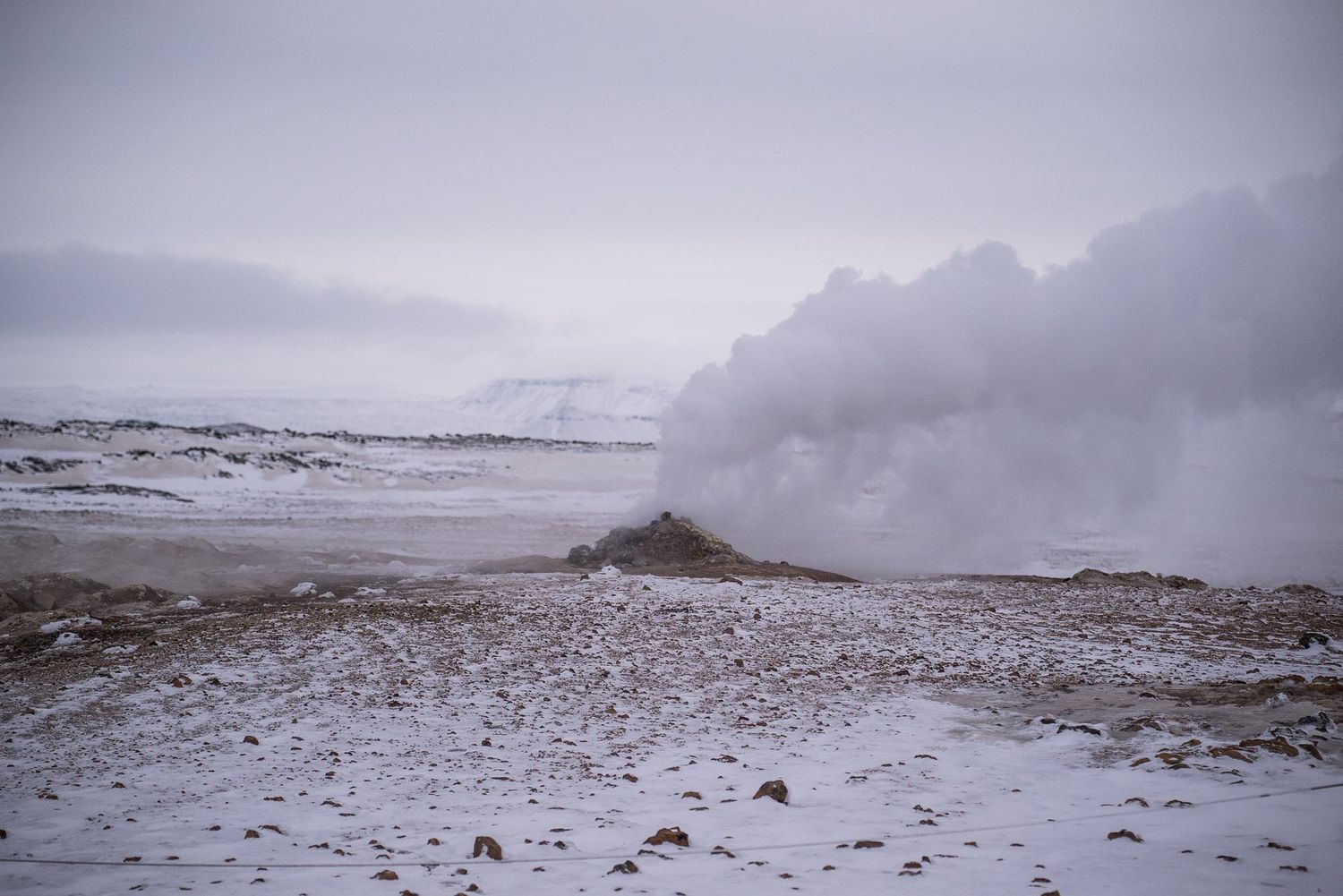 Steam rises from a geothermal hot spring in a snowy winter landscape under cloudy skies in Iceland.