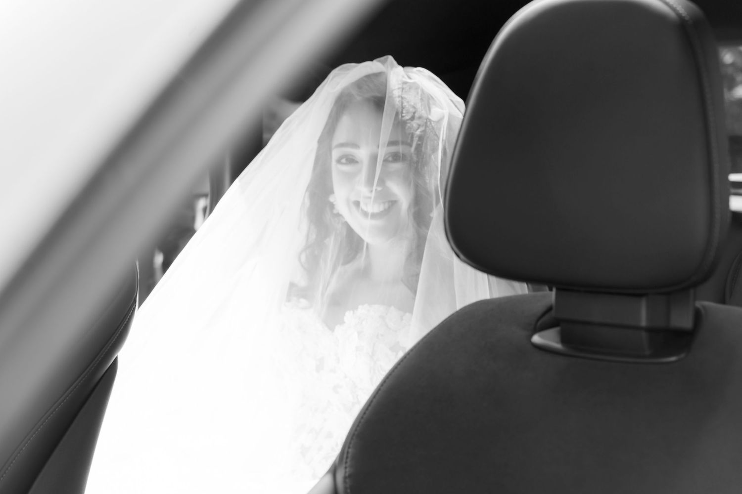 Eine Person in einem weißen Brautschleier sitzt lächelnd in einem Auto, während Sonnenlicht durch das Fenster strömt.