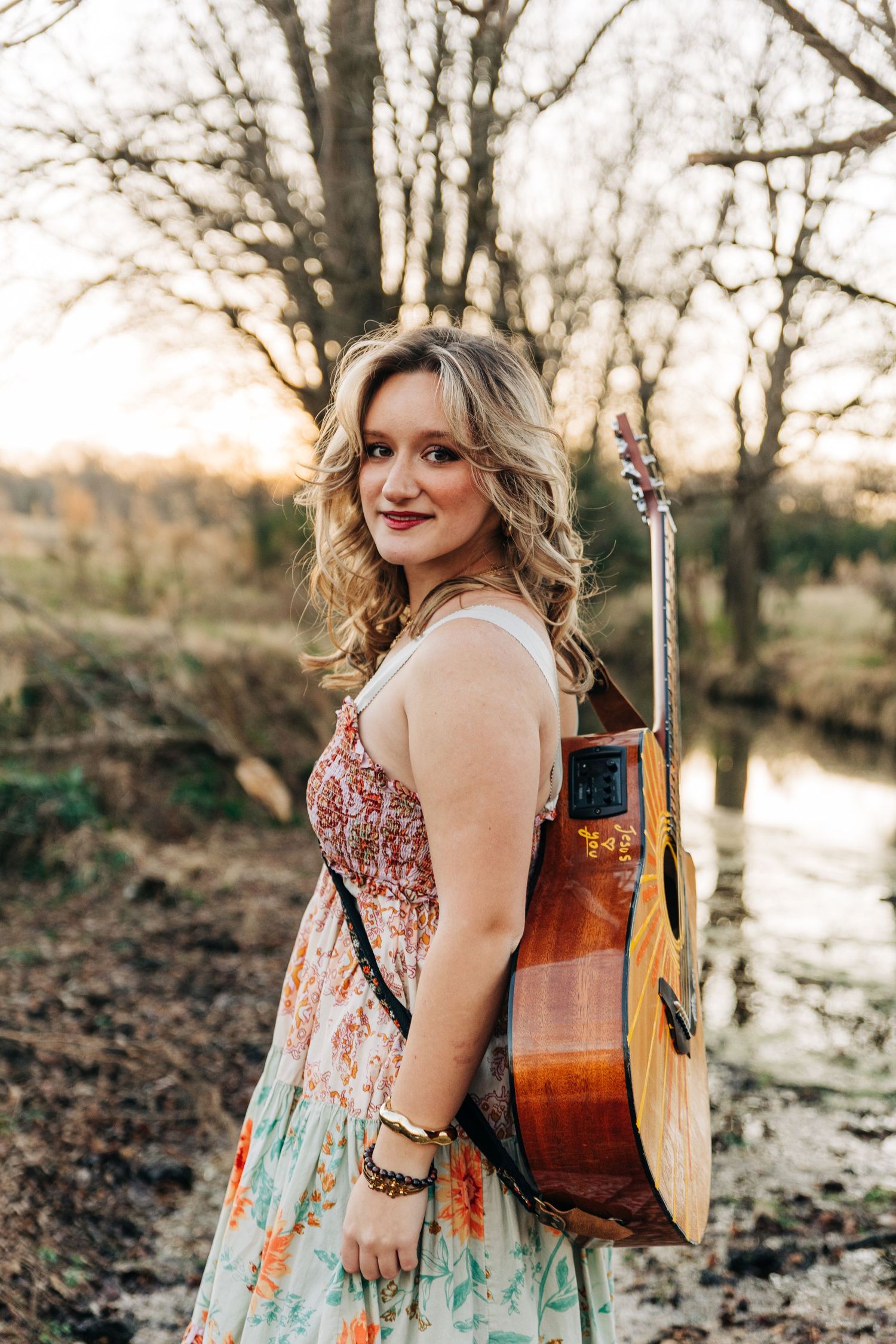 A musician in a floral dress poses with an acoustic guitar by a creek at sunset in a natural outdoor setting.