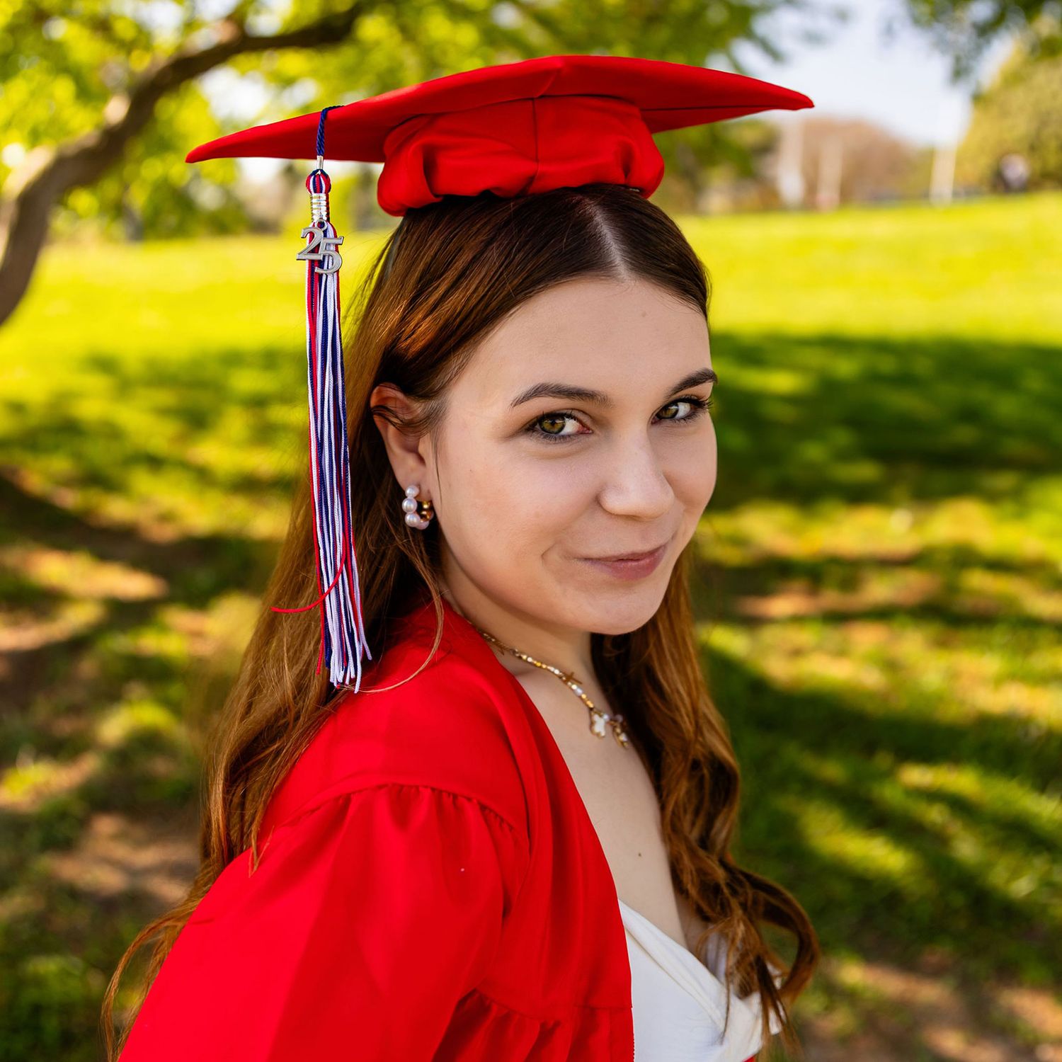 Graduate in red cap and gown poses outdoors on a sunny day with trees and greenery in the background at Memorial Park in Omaha, NE.