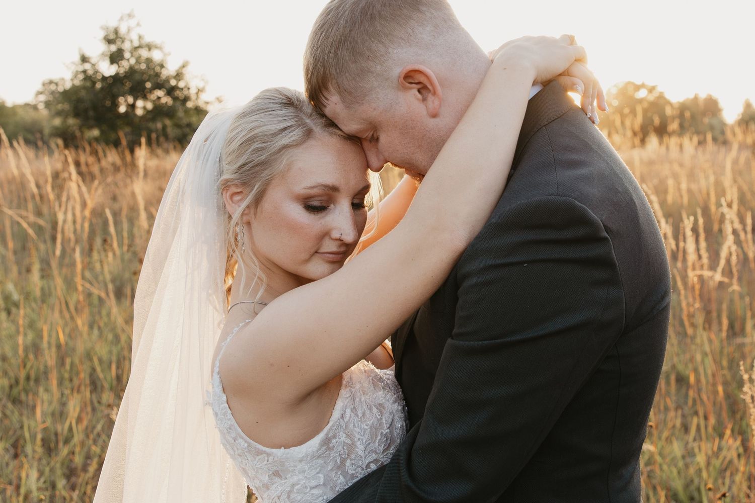 A romantic wedding couple embraces in a golden sunlit field during sunset.