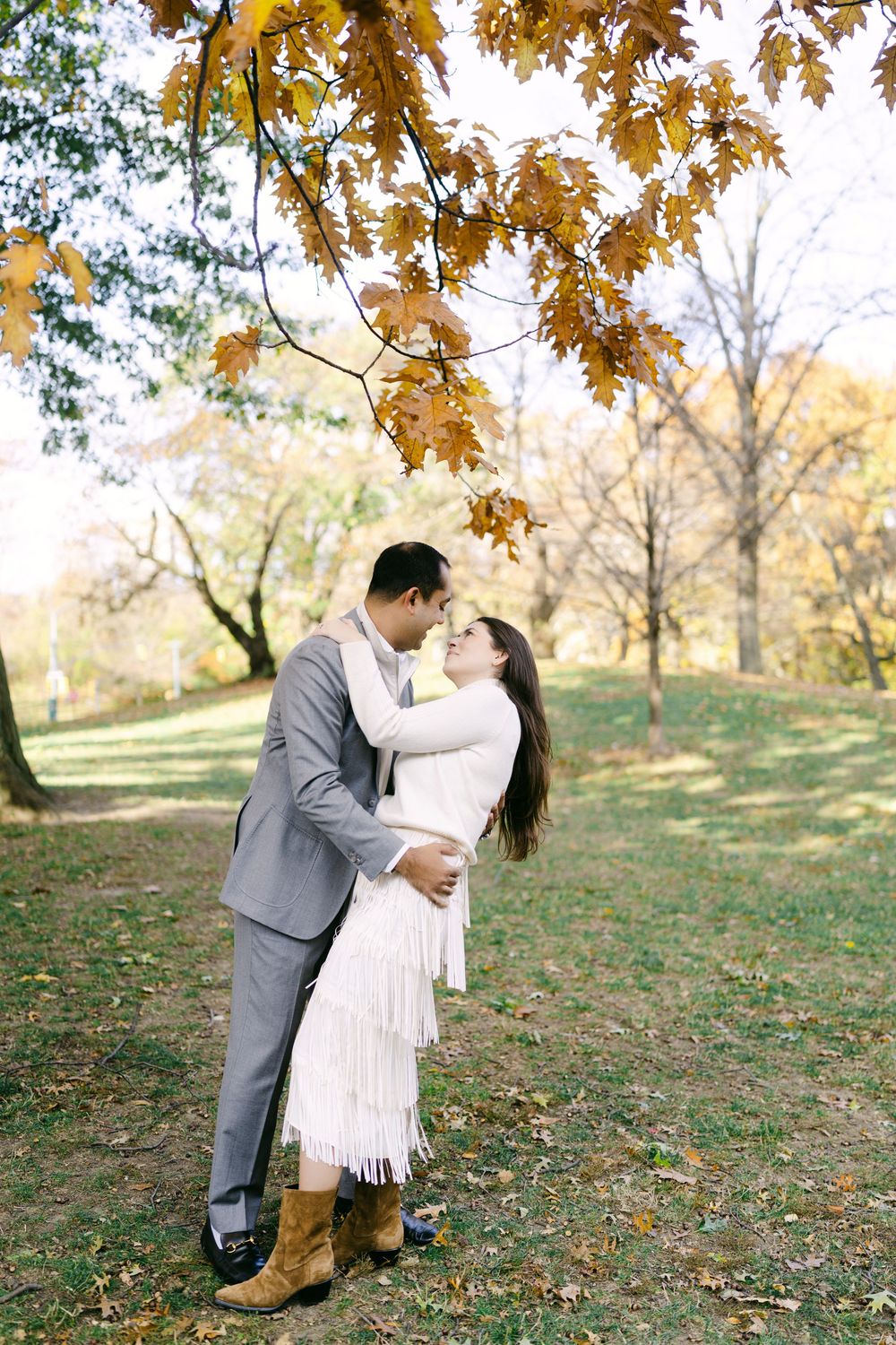 A romantic autumn wedding photo with golden maple leaves overhead as a couple embraces on fallen leaves in a park.