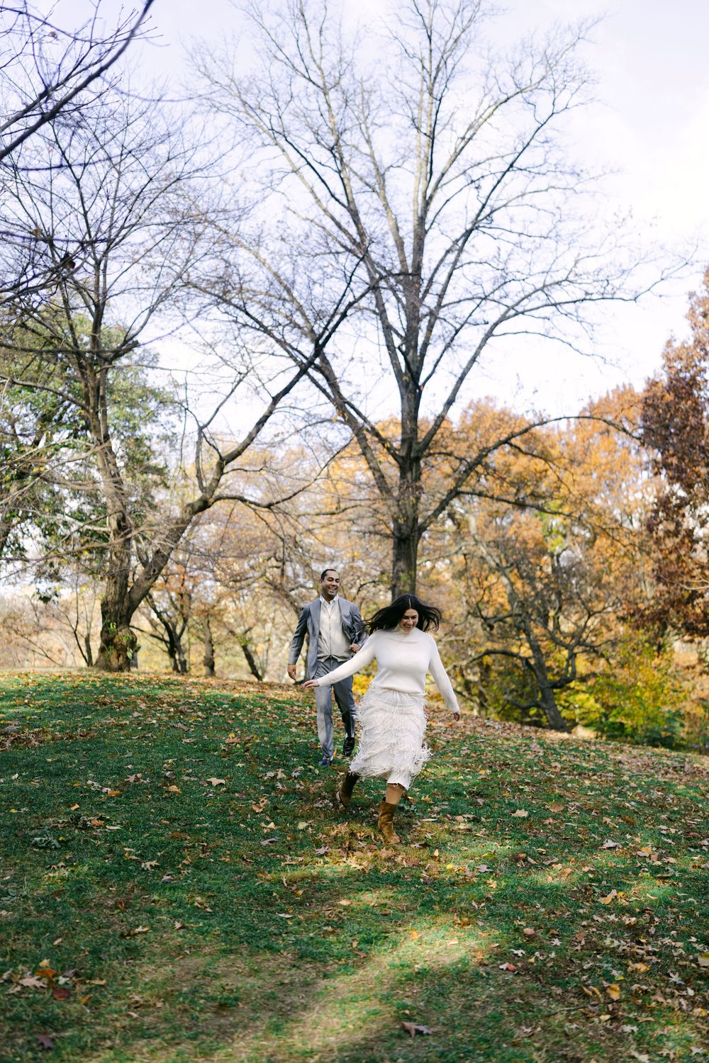 A couple playfully runs and dances on a grassy autumn hillside surrounded by bare trees.