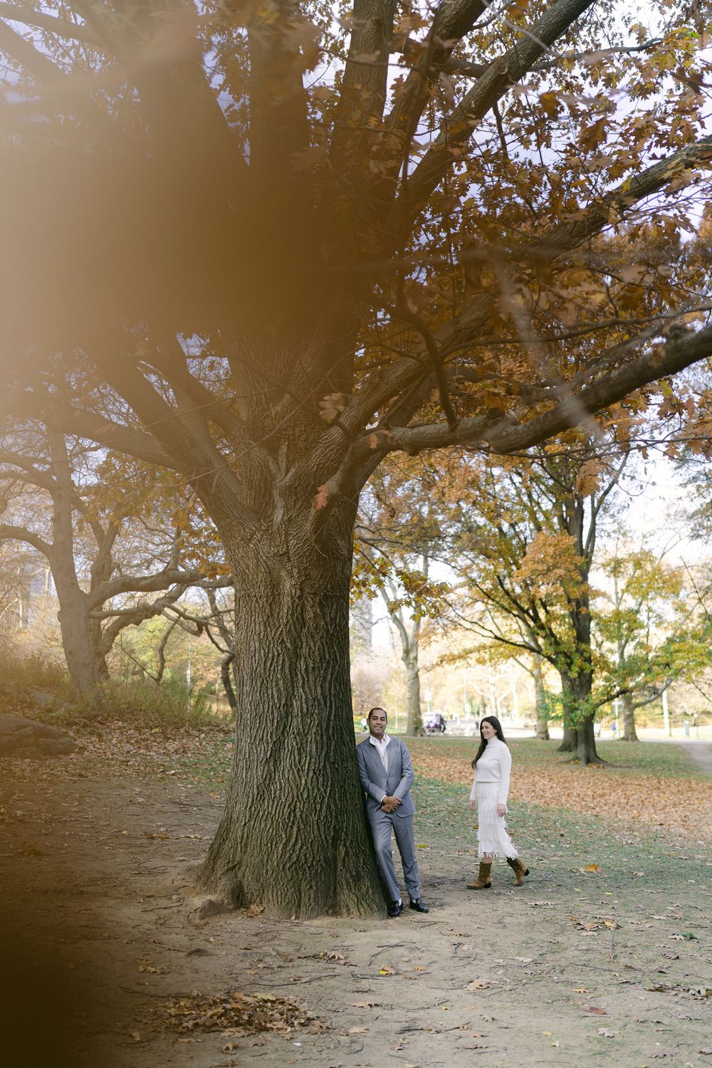 A couple poses playfully beneath large oak trees on an autumn day in a park setting.