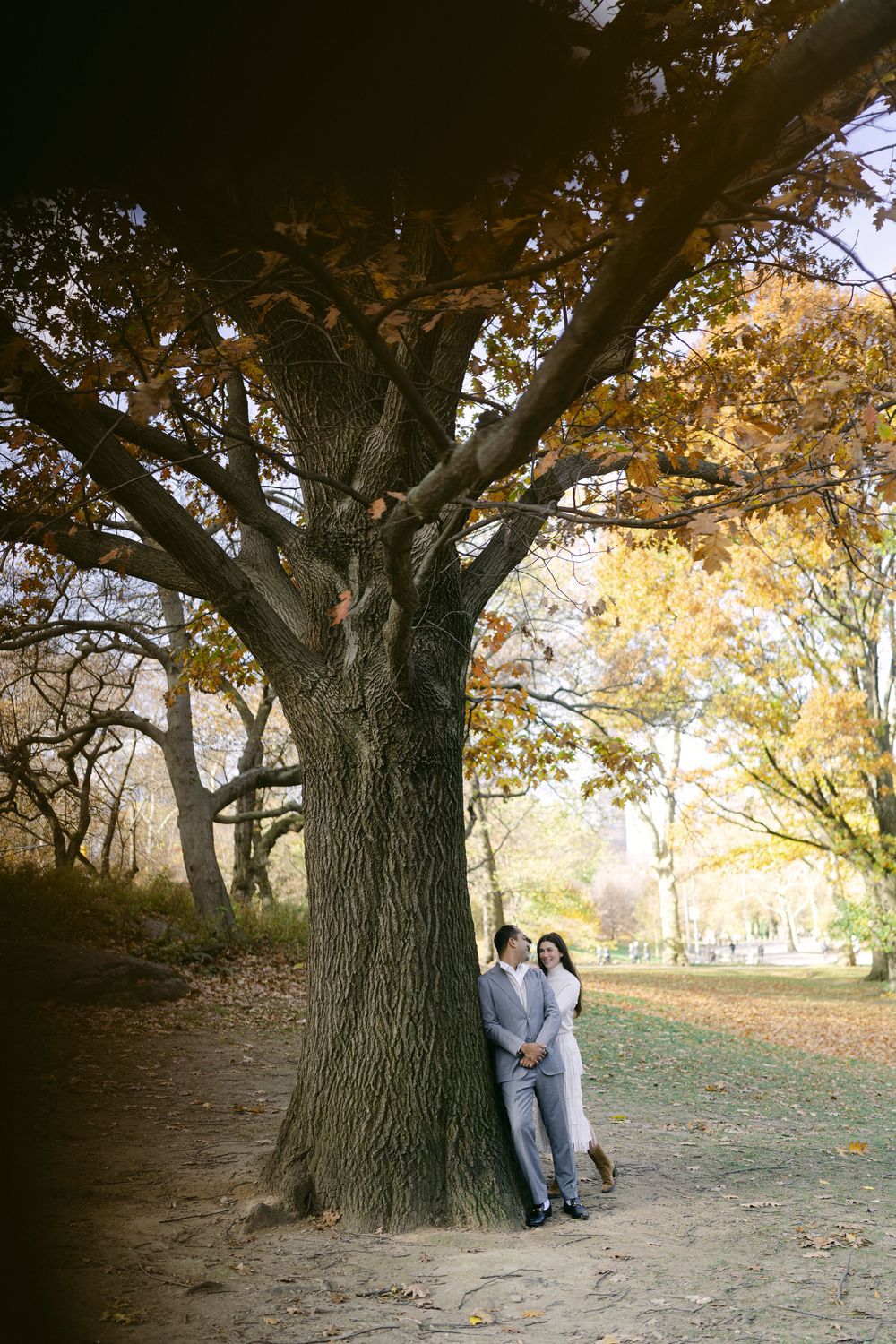 A couple shares an intimate moment against a large tree trunk in an autumn park setting.