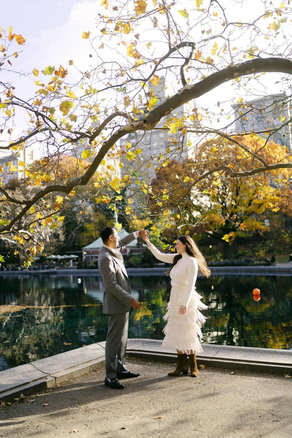 Couple in formal attire dancing and twirling under autumn trees by a serene lake in a park setting.