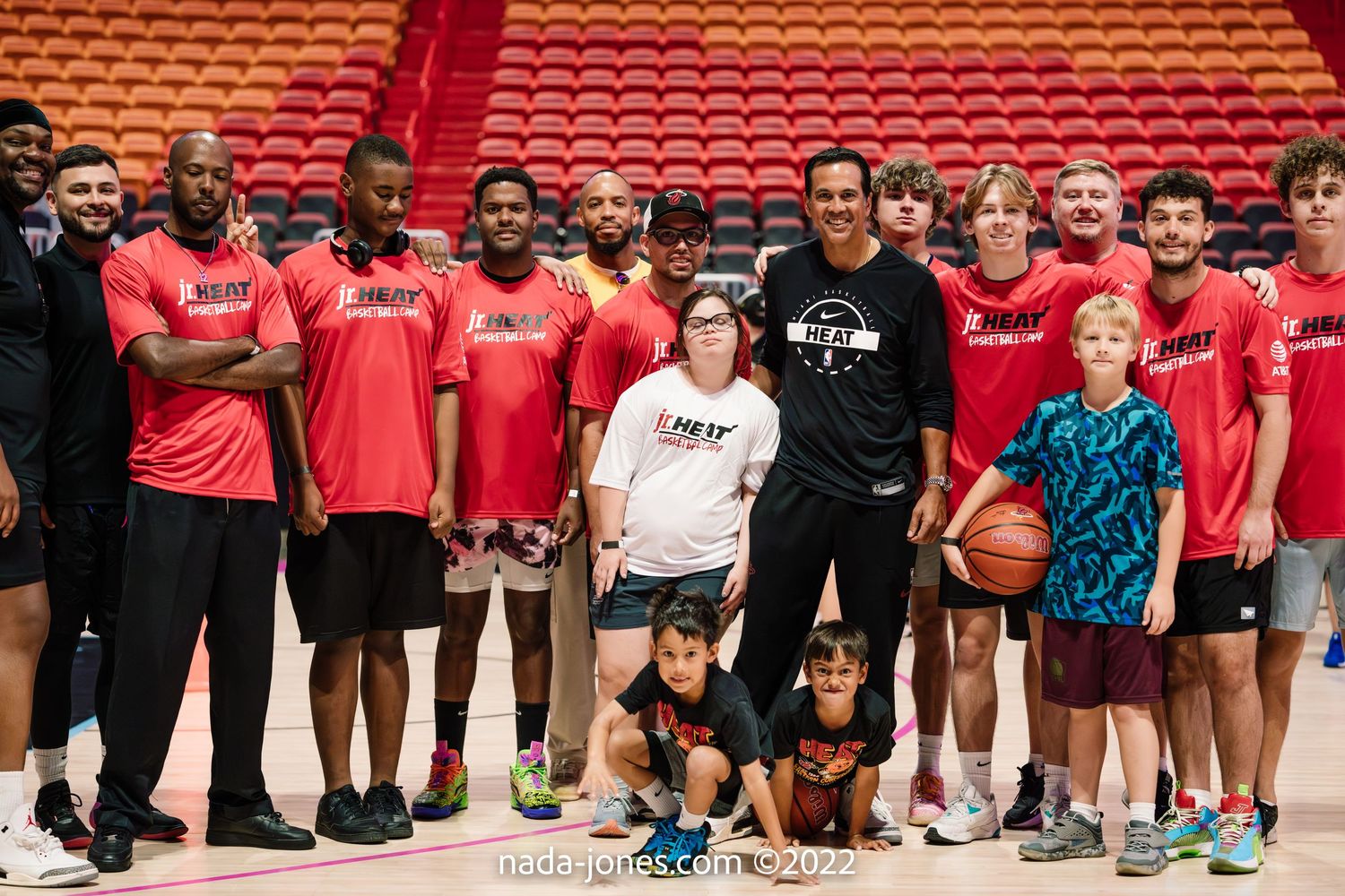 A large group of basketball players and supporters pose together in red shirts on a basketball court.